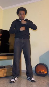 Moletom preto sobre uma camisa branca com colarinho, jeans escuros e ténis pretos com pormenores brancos. Em pé dentro de casa ao lado de uma TV e de uma pequena lareira decorativa.