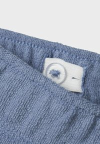 Blaues Frottee-Gewebe mit strukturierten Streifen, ausgestattet mit einem weißen Etikett und einem klaren Knopfverschluss. Nahaufnahme des Stoffs und der Details.