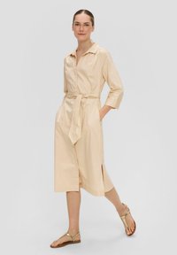 Beige, knielanges Kleid aus Baumwollmischung. Verfügt über einen Kragen, Dreiviertelärmel, geschnittene Taille und Seitenschlitze.