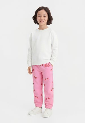 Kind mit mittellangem dunklem Haar, das ein weißes Sweatshirt, rosa Jogginghosen mit Erdbeeraufdruck und "GAP"-Logo sowie weiße Turnschuhe trägt und lächelt.