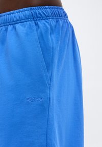 Μπλε σορτς με ελαστική μέση, πλαϊνή τσέπη και κεντημένο λογότυπο "BORG" στο αριστερό μπούτι, που φοριέται από άτομο με σκουρόχρωμο δέρμα.