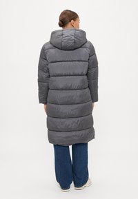 Cappotto lungo grigio imbottito con cappuccio, caratterizzato da sezioni a cucitura orizzontale, abbinato a jeans a gamba larga e sneakers chiare.