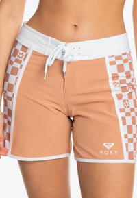 Persikofärgade simshorts med vit midja, rutigt mönster på sidorna, snörningsstängning och Roxy-logotyp. Slät textur och åtsittande design.