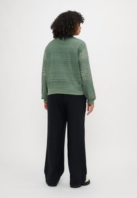 Sweat-shirt vert en crochet avec des poignets et une taille en côte, associé à un pantalon large noir et des chaussures noires épaisses.