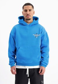 Blauwe katoenen hoodie met een voorzak en grafische tekst. De mouwen en de onderrand zijn geribbeld, en het is gelaagd over een witte T-shirt.