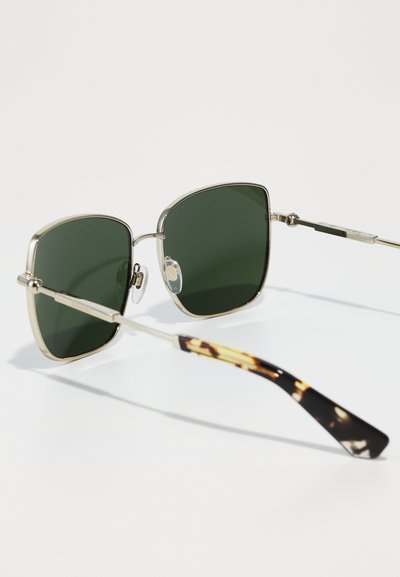 Ochelari de soare pătrați cu lentile verzi închise, montură metalică aurie și brațe cu model de țestoasă. Pernele pentru nas sunt transparente pentru confort.