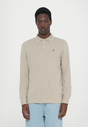 Beige langermet polo-skjorte med krage, kontrasterende hvit kant og liten brodert logo på venstre bryst. Kombinert med lyseblå jeans.