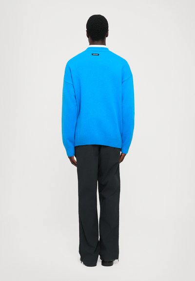 Blauer gestrickter Pullover mit langen Ärmeln, gerippten Bündchen und Saum sowie einem Etikett am Hals. Kombiniert mit schwarzen weit geschnittenen Hosen.