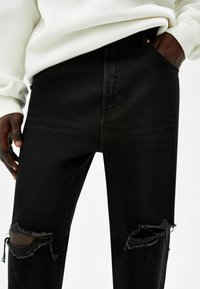 Černé džínové kalhoty z denimu s roztrženými detaily na kolenou, lehce ohlé a úzkým střihem. Kapsy na obou stranách, doplněné krémovou mikinou.