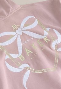 Light pink hoodie featuring white ribbon and heart design with gold accents. "PARIS BAKER by Ted Baker" printed in bold, clear text. Soft texture.