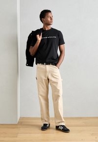 Zwart t-shirt met wit logo, beige broek, zwarte jas over de schouder, zwarte sneakers met witte strepen. Houten vloer, grijze achtergrond.