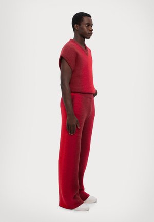 Chaleco de punto rojo y pantalones de pierna ancha hechos de material texturizado. El conjunto es holgado, con un escote en V y bolsillos laterales. Zapatos blancos.