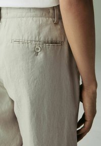 Pantaloni di lino beige chiaro con una tasca posteriore, dotati di chiusura a bottone e dettagli di cucitura sottili. Il tessuto testurizzato è ben visibile.