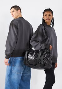 Dva modela nose oversized tamnosive dukserice. Jedan model nosi sjajnu crnu torbu sa kontrastnim trakama. Prikazani su i plavi traperice.