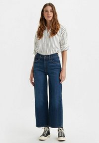 Jean de pierna ancha azul con cintura alta, combinado con una camisa blanca de botones con rayas verticales negras y zapatillas altas negras.