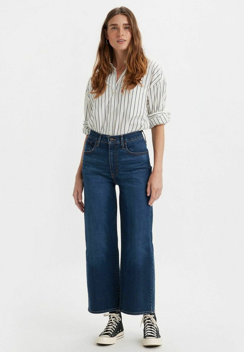 Jean de pierna ancha azul con cintura alta, combinado con una camisa blanca de botones con rayas verticales negras y zapatillas altas negras.