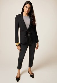 Zwarte op maat gemaakte blazer met een strik in de taille, gedragen over een grijze structuurtop en gecombineerd met donkere jeans en zwarte flats met lintaccenten.