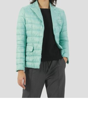 Kvinde iført lyseblå quiltet blazer, sort skjorte og mørkegrå bukser, stående med den ene hånd i lommen.