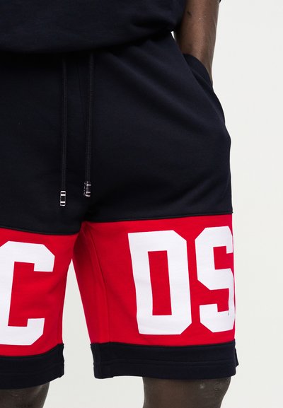 Κοντινό πλάνο μαύρου και κόκκινου σορτς με λευκά γράμματα μπλοκ "C" και "OS" σε κάθε πόδι, που φοριέται από άτομο με τα χέρια στις τσέπες.