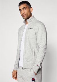 Gray zip-up jacket made of soft fabric, featuring a high collar, side pockets, and a Champion logo on the chest and sleeve.