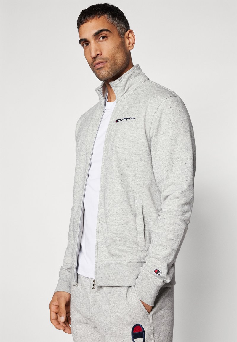 Grijze zip-up jas van zacht materiaal, met een hoge kraag, zijzakken en een Champion-logo op de borst en mouw.
