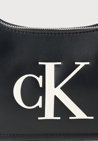 Μαύρη δερμάτινη τσάντα χειρός με εμφανές λευκό λογότυπο "CK", λεία υφή και επάνω κλείσιμο με φερμουάρ. Διακριτικές ραφές λεπτομέρειας.