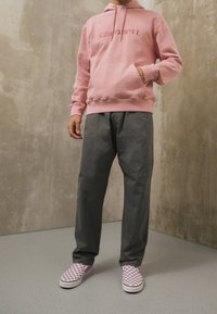 Světlé růžové mikinové tričko s kapucí a přední kapsou, doplněné volnými šedými kalhotami a fialovými slip-on botami s kostkovaným vzorem. Pohodlný a ležérní styl.