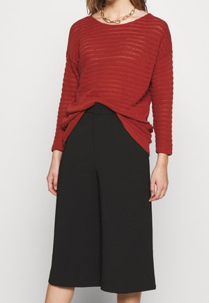 Maglione rosso con texture, dalla vestibilità ampia, maniche lunghe e scollatura rotonda abbinato a pantaloni neri a gamba larga. Collana in oro a catena come accessorio.