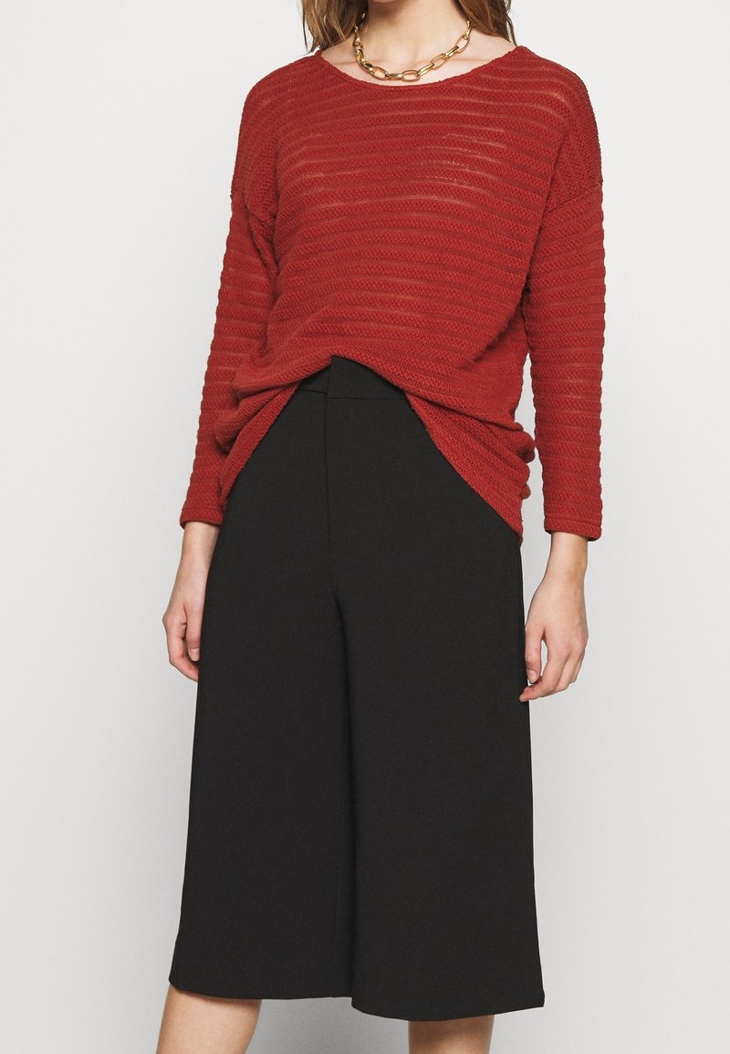 Maglione rosso con texture, dalla vestibilità ampia, maniche lunghe e scollatura rotonda abbinato a pantaloni neri a gamba larga. Collana in oro a catena come accessorio.