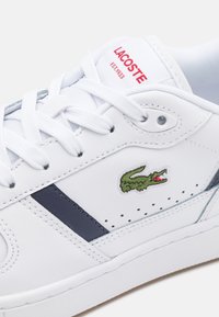 Basket blanche Lacoste avec lacets blancs, bande marine, logo crocodile vert et texte rouge "LACOSTE EST. 1933" sur la languette.
