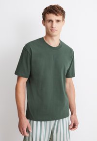 Dunkelgrünes Baumwoll-T-Shirt mit rundem Ausschnitt und kurzen Ärmeln, mit dezent geprägten Buchstaben auf der Vorderseite. Gestreifte Hosen sind sichtbar.