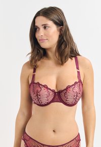 Sostén de encaje burdeos con bordes ondulados, tejido translúcido y bordado floral intrincado. Tirantes ajustables con acentos de hardware en tono dorado.