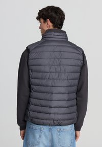 Ciemnoszara kamizelka typu puffer z poziomym pikowaniem, wysokim kołnierzem i lekkim materiałem, noszona na czarnej bluzie z długim rękawem i niebieskich dżinsach.