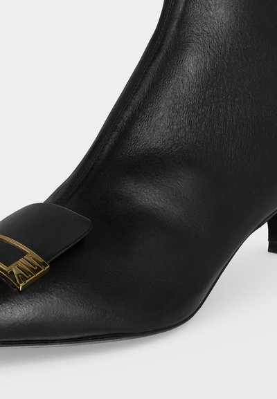 Bottine noire en cuir à bout pointu, talon bas et bride noire décorative avec boucle en métal doré.