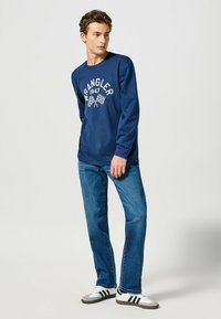 Granatowa koszula z długim rękawem z białym nadrukiem "Wrangler 1947" oraz w szachownicę. W połączeniu z niebieskimi dżinsami i szaro-białymi butami sportowymi.