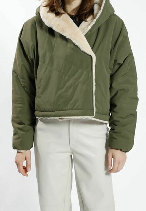 Person trägt eine kurz geschnittene olivgrüne gesteppte Jacke mit beigem Kunstpelzfutter und hellen Hosen vor weißem Hintergrund.