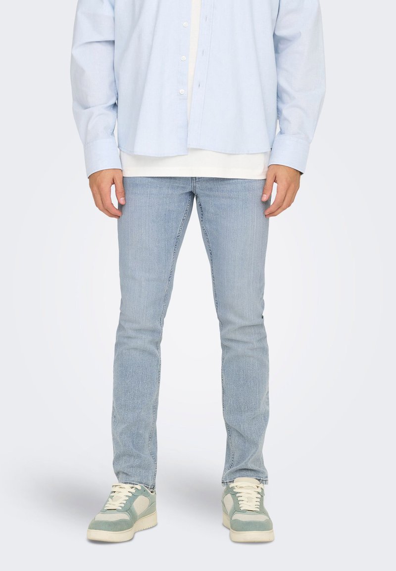 Homme portant un jean bleu clair, des baskets blanches et vertes, et une chemise bleu clair boutonnée sur un t-shirt blanc, debout sur un fond uni.