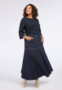 Bluză tricotată de culoare albastră închisă cu mâneci trei sferturi, asortată cu o fustă lungă, evazată din denim, având buzunare și detalii vizibile de cusătură.