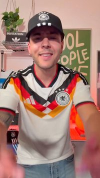 Jeune homme souriant portant un maillot de football d'Allemagne et une casquette noire à l'intérieur, avec une plante et un panneau "Cool People Love" en arrière-plan.