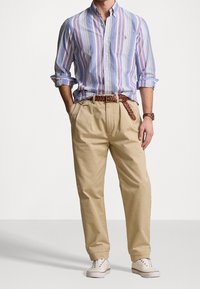 Camisa de botones a rayas en azul, rojo y blanco; pantalones de algodón beige con un cinturón trenzado; zapatos blancos con detalles de ojales.