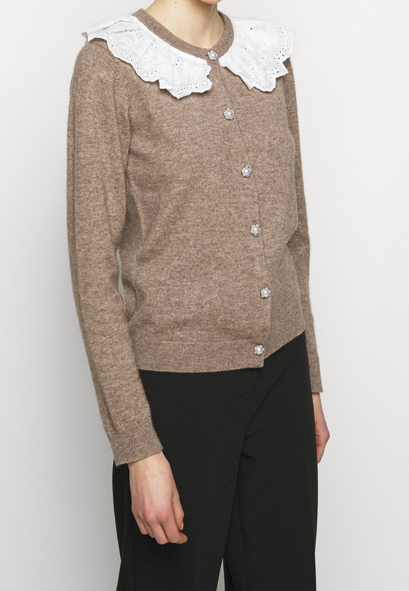 Cardigan tricoté marron clair avec boutons en perle et grand col en dentelle blanche, porté avec un pantalon noir sur une personne debout.