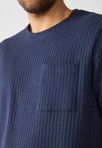 Pulover tricotat texturat de culoare bleu marin, cu gât rotund și un buzunar pătrat pe piept. Dispune de un model în formă de vafe și este realizat dintr-un material moale și respirabil.