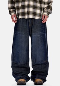 Άτομο που φοράει φαρδιά σκούρα μπλε τζιν, καφέ παλιά μπότες και πουκάμισο με καρό μοτίβο σε μπεζ και πράσινο με μανσέτες από ορείχαλκο.