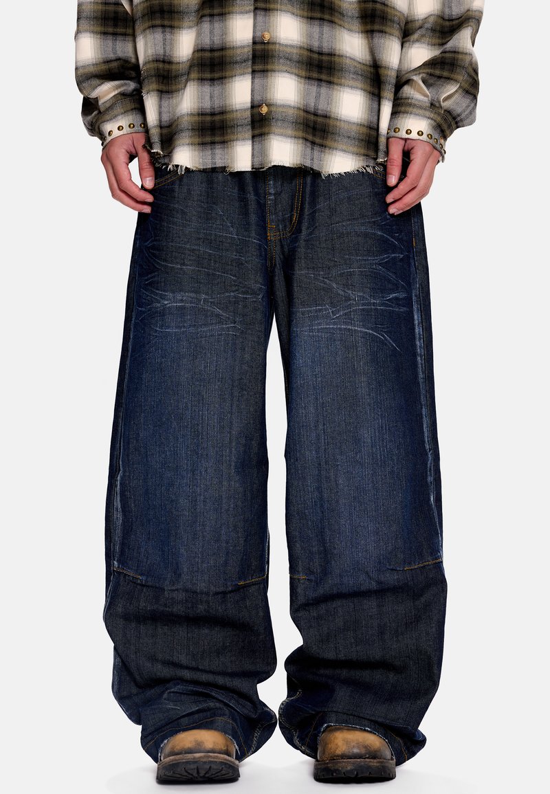 Άτομο που φοράει φαρδιά σκούρα μπλε τζιν, καφέ παλιά μπότες και πουκάμισο με καρό μοτίβο σε μπεζ και πράσινο με μανσέτες από ορείχαλκο.