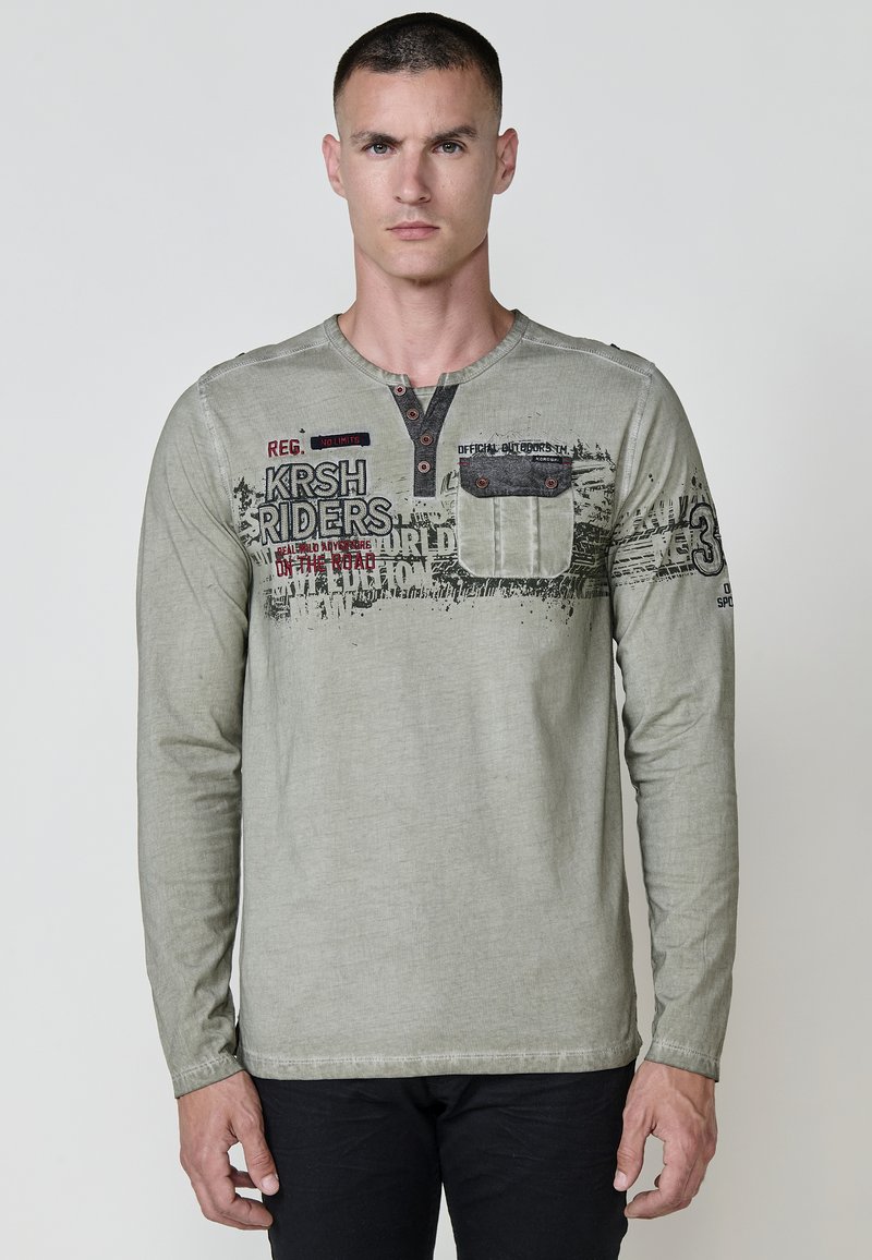 Hosszú ujjú olajzöld ing puha anyagból, henley nyakkivágással, mellzsebkérdéssel, merész grafikákkal és fekete, vörös kontrasztos részletekkel.