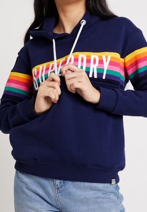 Femme ajustant les cordons blancs sur un sweat à capuche bleu marine avec des bandes horizontales arc-en-ciel et le texte "Superdry", portant un jean bleu clair.