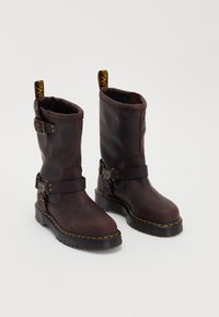 Brown leather boots with a mid-calf design, featuring a strap and buckle, a thick black sole, and yellow stitching accents.