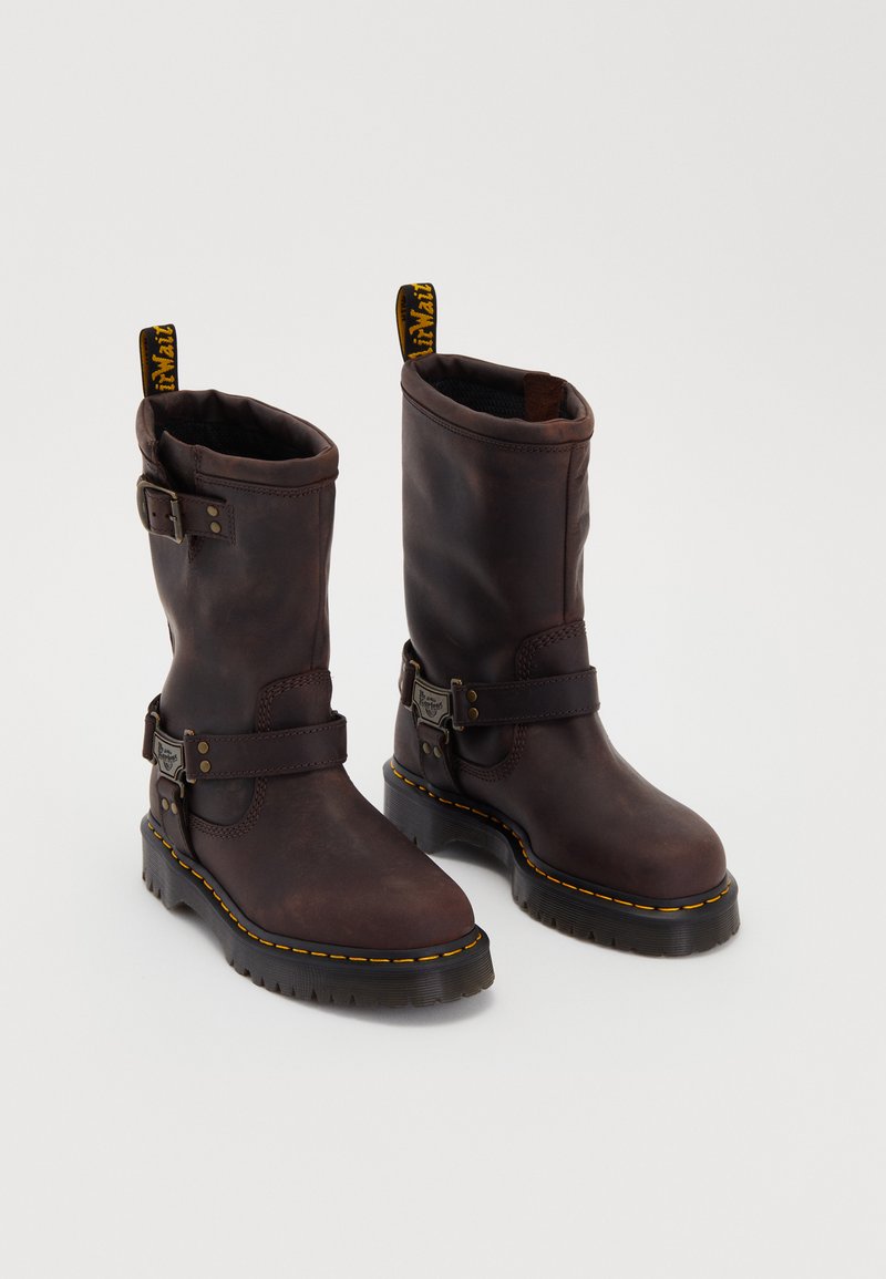Brown leather boots with a mid-calf design, featuring a strap and buckle, a thick black sole, and yellow stitching accents.