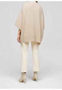 Poncho beige lavorato a maglia con motivo testurizzato, vestibilità oversize e bordi a costine, abbinato a pantaloni a campana color crema e stivali beige alla caviglia.