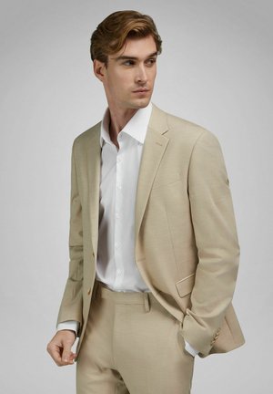 Mann in beigem Anzug und weißem Hemd, der nach links schaut, eine Hand in der Tasche und die andere entspannt an der Seite.